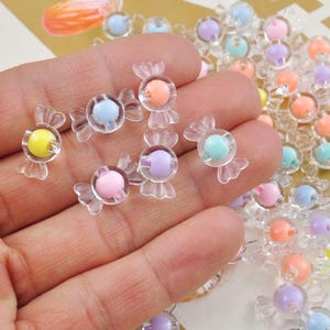 May include: Assortment of colorful candy-shaped resin charms with clear plastic wrappers. The charms are in various colors, including pink, blue, yellow, and purple.