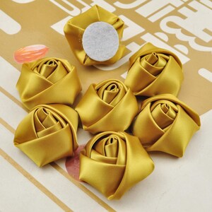 May include: A set of seven gold satin fabric rose flower embellishments. Each flower is approximately 2 inches in diameter and has a flat back with a white adhesive circle.
