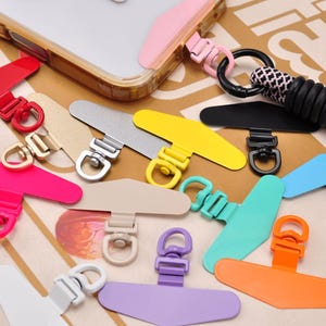 Può includere: Una collezione di colorati patch per cordino per telefono in varie tonalità, tra cui rosso, rosa, giallo, argento, nero, blu, verde, arancione, viola e beige. Questi accessori sono progettati per essere attaccati a una custodia del telefono, con un anello per un cordino.