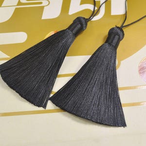 May include: Two black tassel ornaments with a long, thin, fringed design. The tassels are attached to a thin black cord.