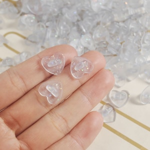 May include: A close-up of a hand holding four clear heart-shaped buttons with iridescent glitter. The buttons have a small hole in the center for sewing.