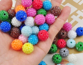 May include: A hand holding a variety of colourful, round, plastic beads. The beads are covered in small, clear rhinestones. The beads are approximately 2.5 cm in diameter.