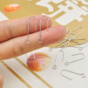 May include: A collection of silver-colored earring hooks. Two hooks are held between fingers, with several more scattered on a patterned surface. Each hook has a looped bottom and a curved top.