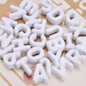 Könnte beinhalten: Eine Ansammlung weißer Kunststoff-Alphabetperlen mit verschiedenen Buchstaben in einer abgerundeten Blockschrift. Jede Perle hat eine glatte, glänzende Oberfläche und ein Loch in der Mitte zum Auffädeln, lose verstreut.