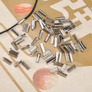 Puede incluir: Una colección de pequeñas cuentas de crimpado plateadas esparcidas sobre una superficie estampada. Las cuentas son rectangulares con un borde crimpado, diseñadas para la fabricación de joyas. También se ve un cordón negro con un extremo plateado.