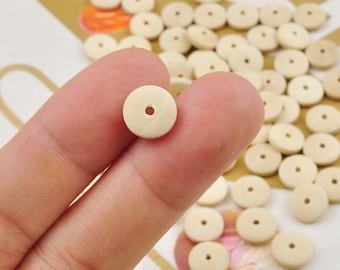May include: A collection of small, round, light beige wooden beads with a central hole. One bead is held between fingers, with many more scattered in the background. These beads are suitable for jewelry making and craft projects.