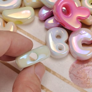 May include: A collection of iridescent, glossy, rounded beads in various pastel colours including pink, cream, and lavender. The beads feature smooth, organic shapes with central holes for threading, showcasing a reflective, pearlescent finish.