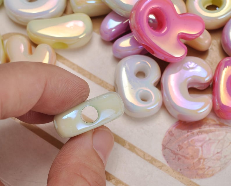 May include: A collection of iridescent, glossy, rounded beads in various pastel colours including pink, cream, and lavender. The beads feature smooth, organic shapes with central holes for threading, showcasing a reflective, pearlescent finish.