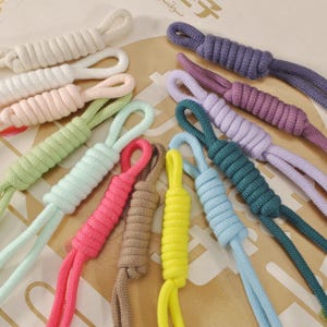 Peut inclure: Une collection de cordons tressés colorés dans diverses teintes, notamment rose, vert, bleu et violet. Chaque cordon a une extrémité en boucle et un nœud cylindrique décoratif. Les cordons sont disposés sur une surface à motifs.