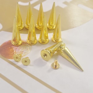 May include: A set of ten gold-colored metal pyramid-shaped studs with screws. The studs are about 1 inch tall and have a pointed tip. The screws are about 1/2 inch long and have a Phillips head.