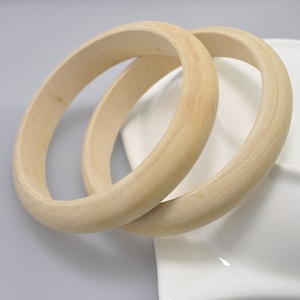 Puede incluir: Dos brazaletes de madera de color claro, con un diseño liso y redondeado. Los brazaletes están hechos de madera natural y descansan sobre una superficie blanca. Los brazaletes son un accesorio simple pero elegante.