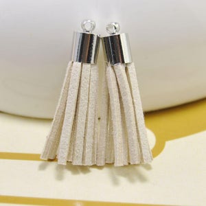 Puede incluir: Dos borlas de ante de color blanco roto con tapas de metal plateado. Las borlas miden aproximadamente 5 cm de largo.