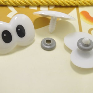 May include: A collection of white plastic craft supplies, including a pair of googly eyes with black pupils, a mushroom-shaped piece, and several other small, round components. The items are arranged on a light yellow surface.