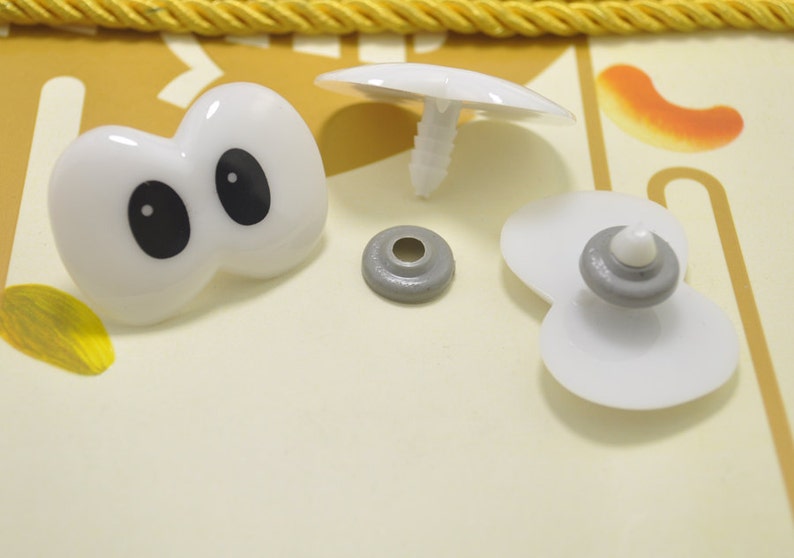 May include: A collection of white plastic craft supplies, including a pair of googly eyes with black pupils, a mushroom-shaped piece, and several other small, round components. The items are arranged on a light yellow surface.