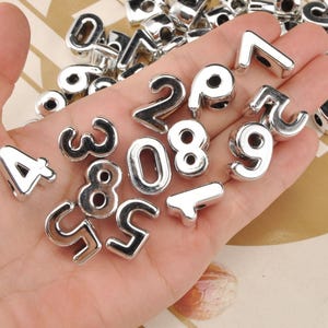 Puede incluir: Una colección de cuentas de números de metal plateado brillante, que incluyen los números del 0 al 9, se muestra en una mano. Las cuentas tienen una superficie lisa y reflectante y están diseñadas con un agujero para ensartar.