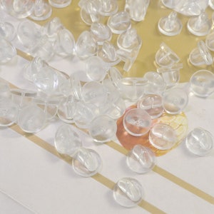 Könnte beinhalten: Eine große Menge an transparenten Kunststoff-Ohrring-Rücken. Die Rücken sind rund mit einem kleinen Stift in der Mitte.
