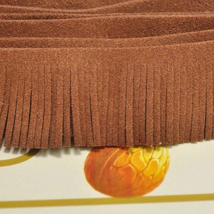 Puede incluir: Primer plano de flecos de ante marrón. Los flecos están cortados en tiras largas y finas, creando un efecto texturizado y en capas. El material parece suave y flexible. La imagen sugiere un producto artesanal o textil.