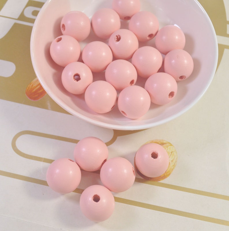 May include: A bowl filled with pink wooden beads. The beads are round and have a hole in the center. There are also a few beads scattered on a white surface.