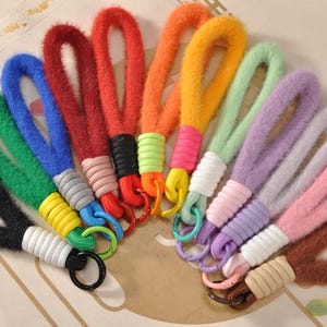 May include: A collection of colorful wrist straps in various shades, including black, green, blue, red, orange, yellow, and purple. Each strap features a soft, fuzzy texture and is attached to a metal ring. The straps are arranged in a circular pattern.