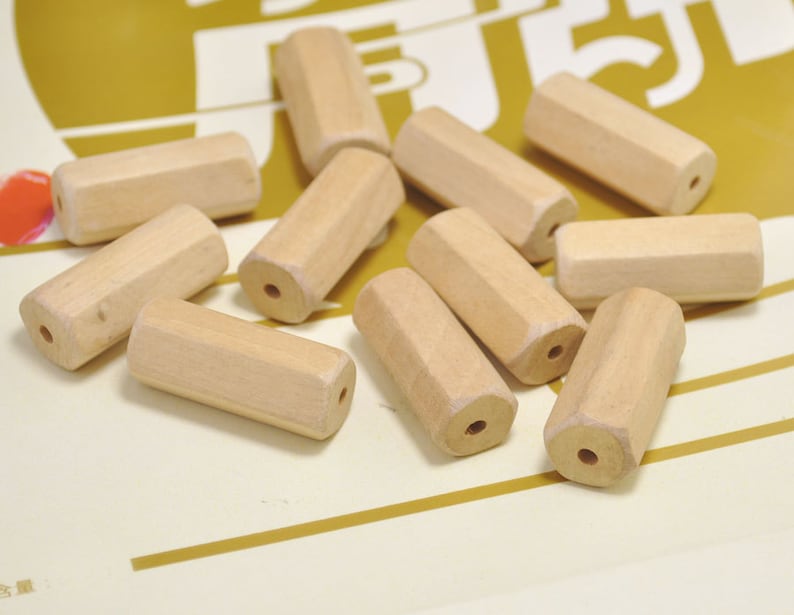 May include: A group of 10 natural wood beads, each with a hexagonal shape and a hole drilled through the center. The beads are scattered on a yellow surface.