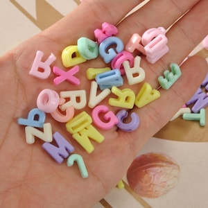 May include: A selection of pastel-coloured alphabet beads in various shapes and sizes. The beads are in shades of pink, yellow, blue, green, and purple. The letters are scattered across a hand, suggesting their use in crafting or jewellery making.