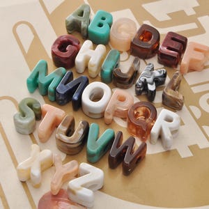 May include: A collection of colorful, marbled resin alphabet letters. The letters are in various colors, including green, brown, white, and peach. The letters are arranged on a light brown surface.