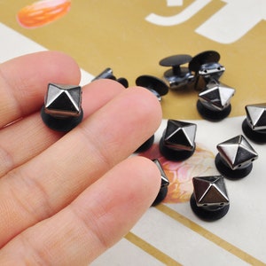 May include: A close-up of a hand holding a black pyramid-shaped stud with a black base. The stud is one of many similar studs scattered on a white surface.