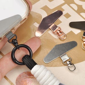 以下が含まれることがあります： シルバー、ローズゴールド、ブラックのメタリック仕上げの携帯電話用ランヤードアタッチメントのクローズアップ。1つは、黒いリングと白と黒のランヤードが付いた携帯電話ケースに接続されています。金属製のアタッチメント。