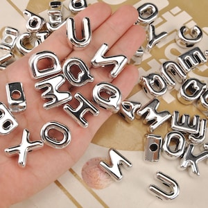 Könnte beinhalten: Silberne Metall-Alphabetperlen in verschiedenen Buchstaben, darunter U, Q und O. Die Perlen sind auf einer Oberfläche verstreut, einige werden in einer Hand gehalten. Die Perlen sind glänzend und haben eine abgerundete, blockartige Schrift.