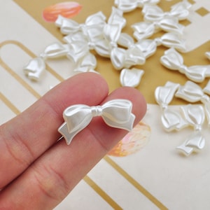May include: A collection of white pearl-like plastic bows. The bows are approximately 2.5 cm in size and have a 3D design.