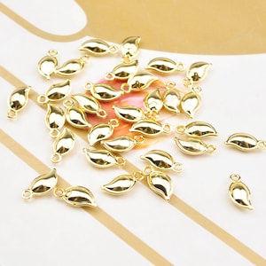 May include: A selection of small, gold-coloured leaf charms. Each charm has a smooth, reflective surface and a small loop at the top for attaching to jewellery or other crafts. The charms are scattered across a light-coloured surface.