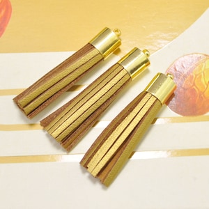 Könnte beinhalten: Drei goldene Lederquasten mit einem goldenen Metallkopf. Die Quasten sind braun und gold.