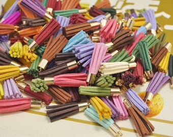 Puede incluir: Una colección de borlas de gamuza de colores con tapas de metal dorado. Las borlas están dispuestas en una pila sobre una superficie blanca.