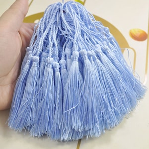 Puede incluir: Un montón de borlas de color azul claro, hechas de hilo suave y sedoso. Las borlas están todas atadas en la parte superior, creando un montón esponjoso y decorativo.