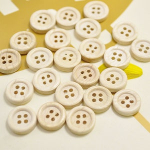 Puede incluir: Una colección de botones de madera natural con cuatro agujeros cada uno. Los botones están dispuestos en un patrón aleatorio sobre una superficie blanca.