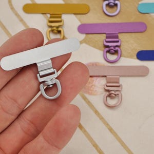 K&ouml;nnte beinhalten: Ein silberner Handy-Anh&auml;nger mit rechteckiger Oberseite und kleinem Ring wird in einer Hand gehalten. Weitere Anh&auml;nger in Gold, Lila, Rosa und Blau sind im Hintergrund zu sehen. Diese Anh&auml;nger sind f&uuml;r Handyh&uuml;llen konzipiert.