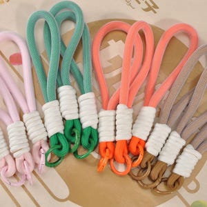 May include: A collection of colorful rope keychains in pink, green, orange, and tan. Each keychain features a loop of thick, woven rope, a white wrapped section, and a metal ring. The keychains are arranged on a patterned surface.