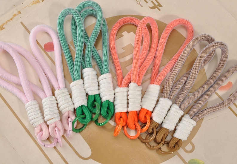 May include: A collection of colorful rope keychains in pink, green, orange, and tan. Each keychain features a loop of thick, woven rope, a white wrapped section, and a metal ring. The keychains are arranged on a patterned surface.