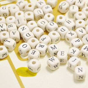 Op de afbeelding: Een verzameling witte houten letter kralen met zwarte letters. De kralen zijn verspreid over een licht beige oppervlak.