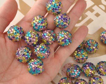 May include: A selection of multi-coloured, round beads. Each bead is covered in small, sparkling rhinestones in shades of blue, pink, yellow, and purple. The beads are held in a hand, showing their size and detail. These beads are suitable for jewellery making.