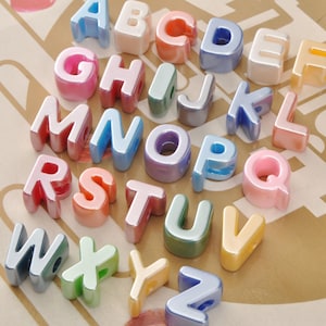 Può includere: Una collezione di perline alfabetiche colorate e lucide. Ogni lettera è di un colore diverso, tra cui rosa, blu, arancione e verde. Le perline sono disposte su una superficie marrone chiaro.