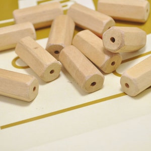 May include: A group of wooden beads in a light brown color. The beads are cylindrical and have a hole through the center. The beads are scattered on a white surface.
