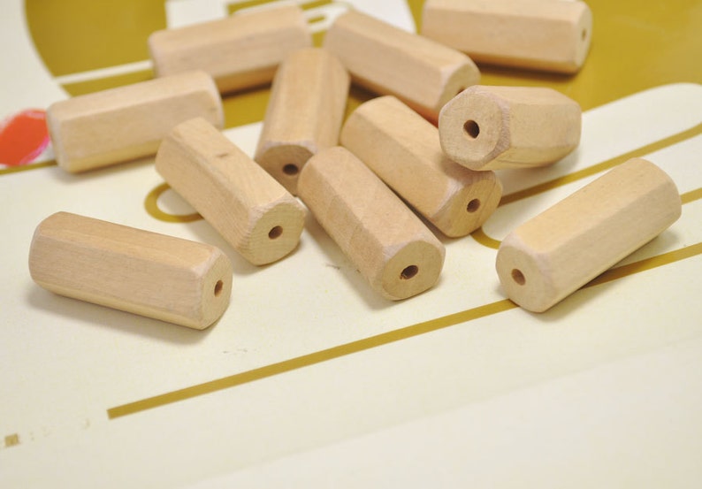May include: A group of wooden beads in a light brown color. The beads are cylindrical and have a hole through the center. The beads are scattered on a white surface.