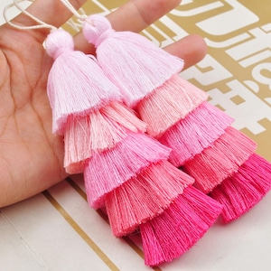 Puede incluir: Dos borlas de flecos de color rosa y blanco degradado con múltiples capas de flecos. Las borlas están atadas con un cordón blanco.