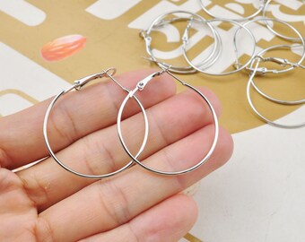 May include: Two silver hoop earrings with a hinged clasp. The earrings are shown on a hand.
