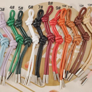 May include: A collection of colorful leather cords with knotted details and silver-toned metal end caps. The cords come in various colors, including silver, blue, green, white, red, and brown. These are likely used for crafting or jewelry making.