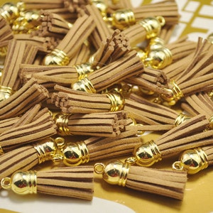 Puede incluir: Un montón de borlas de ante marrón con tapas de metal dorado. Las borlas miden aproximadamente 2,5 cm de largo.