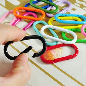 May include: A collection of colorful metal key rings in various shapes and sizes, including a black key ring held in a hand.