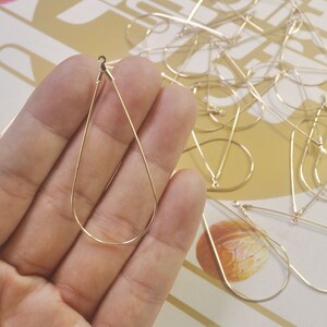 May include: A close-up of a gold teardrop-shaped earring finding. The finding is made of metal and has a smooth finish. The finding is held in a hand.