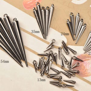 Può includere: Una collezione di ciondoli a forma di cono in metallo argentato con piccoli anelli per la creazione di gioielli. Le dimensioni variano da 1,3 cm a 5,4 cm di lunghezza. I ciondoli sono disposti in gruppi e singolarmente.